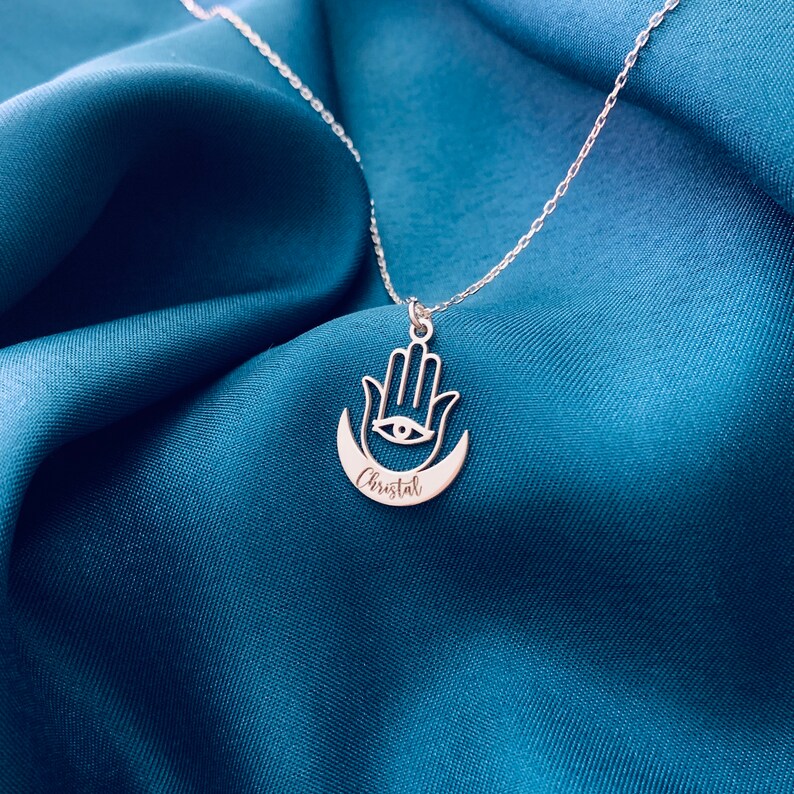 Pode incluir: Colar de prata com um pingente de m&atilde;o de hamsa e um pingente de lua crescente. O pingente tem um olho no centro e o nome "Cristal" gravado na lua.