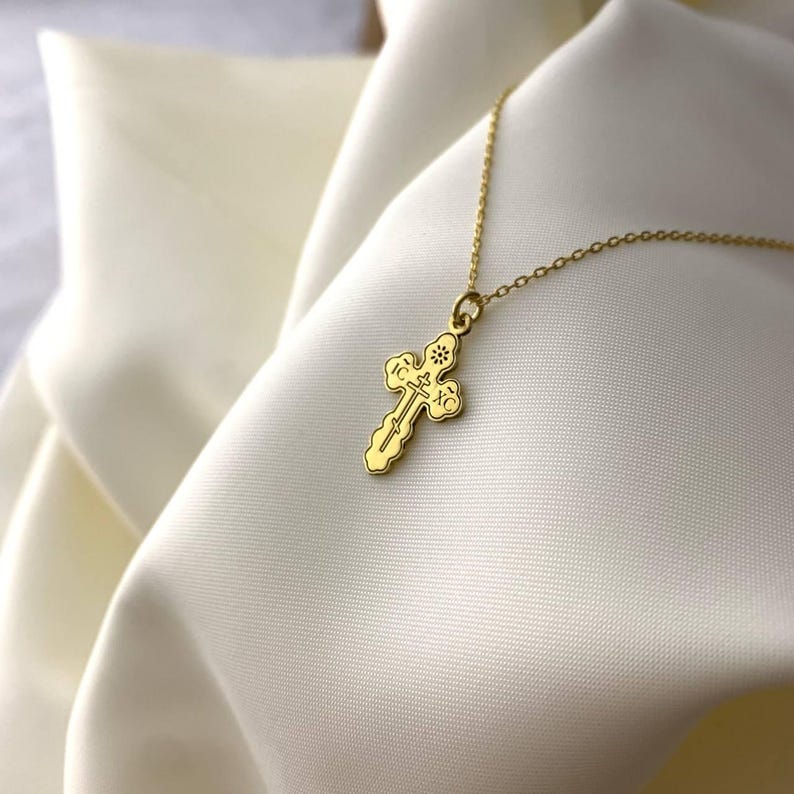 May include: A gold chain necklace with a small, ornate gold cross pendant. The cross has intricate details and is set against a white background.