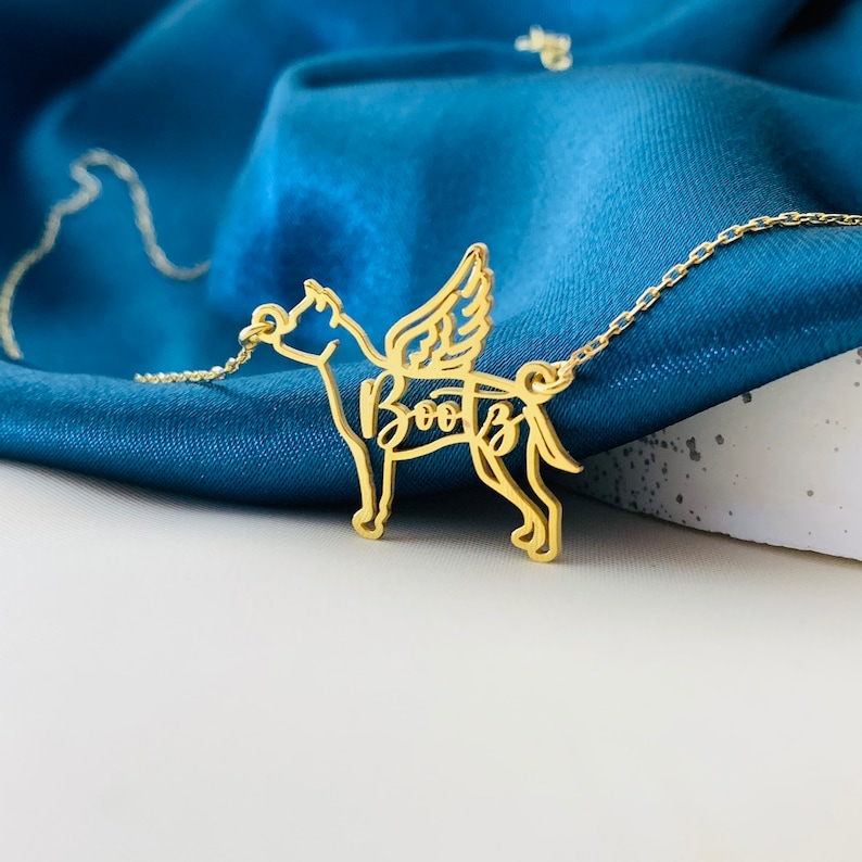 May include: Gold necklace with a dog silhouette pendant. The dog has wings and the name "Boots" is written inside the silhouette.