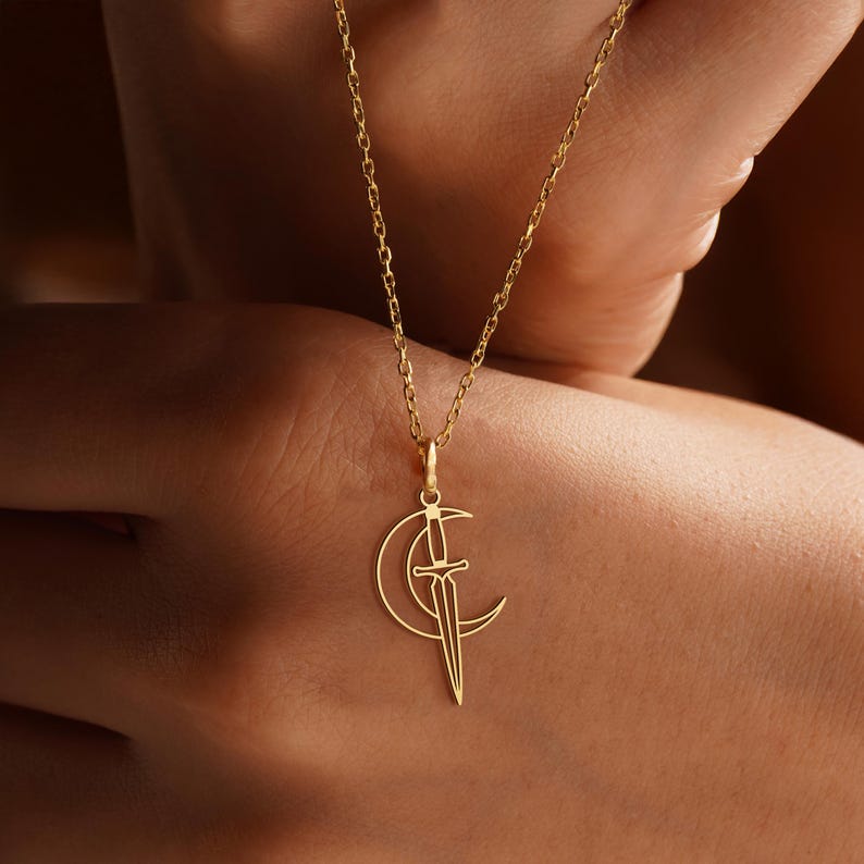 May include: A gold-colored necklace featuring a crescent moon and sword pendant. The pendant is a delicate, open-work design. The chain is a fine link style. The necklace is displayed against a blurred background.