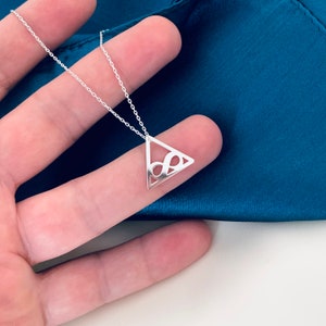 Pode incluir: Um colar de prata com um pingente triangular com um s&iacute;mbolo de infinito vazado. O pingente pende de uma corrente fina. O colar &eacute; exibido em um fundo azul-petr&oacute;leo.