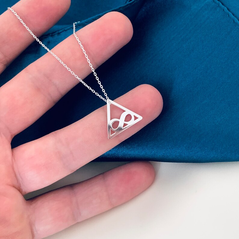 Pode incluir: Um colar de prata com um pingente triangular com um s&iacute;mbolo de infinito vazado. O pingente pende de uma corrente fina. O colar &eacute; exibido em um fundo azul-petr&oacute;leo.