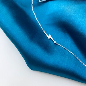 Puede incluir: Un collar de cadena de plata con un colgante de rayo en un fondo de tela azul turquesa.