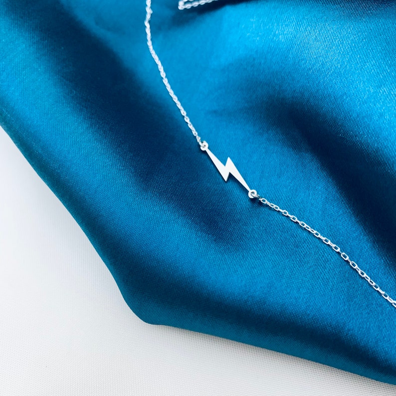Puede incluir: Un collar de cadena de plata con un colgante de rayo en un fondo de tela azul turquesa.