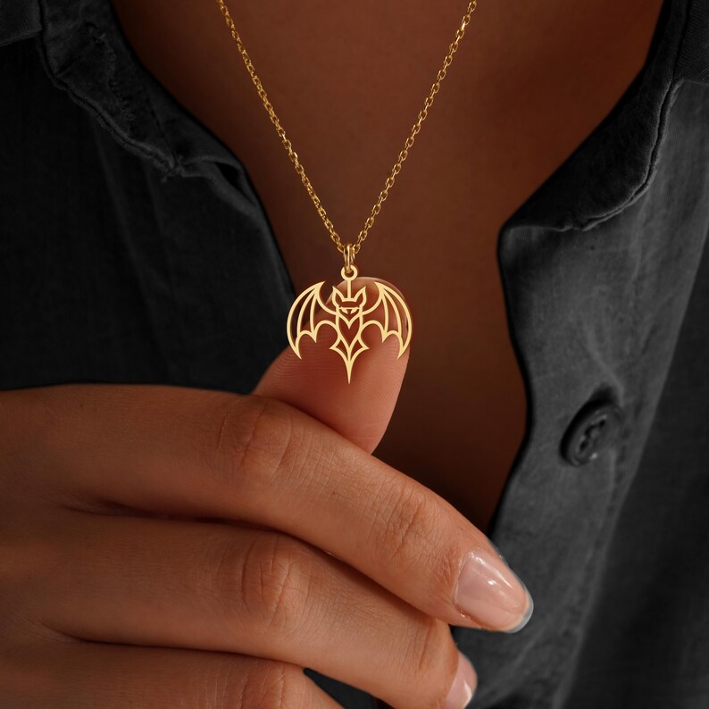 K&ouml;nnte beinhalten: Eine goldfarbene Halskette mit Anh&auml;nger, die ein Fledermaus-Design zeigt. Der Fledermaus-Anh&auml;nger ist aufwendig ausgeschnitten und zeigt die Fl&uuml;gel- und K&ouml;rperdetails. Die Halskette wird vor einem dunklen Hintergrund getragen.