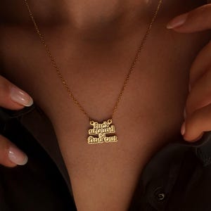 May include: Gold-colored necklace with the phrase "fuck around & find out" in a stylized font. The pendant hangs from a delicate chain. The necklace is displayed against a dark background.