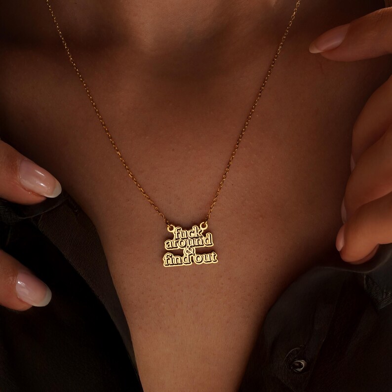 May include: Gold-colored necklace with the phrase "fuck around & find out" in a stylized font. The pendant hangs from a delicate chain. The necklace is displayed against a dark background.