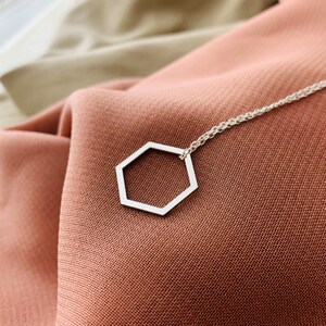 May include: A silver necklace with a hexagon pendant on a brown fabric background.