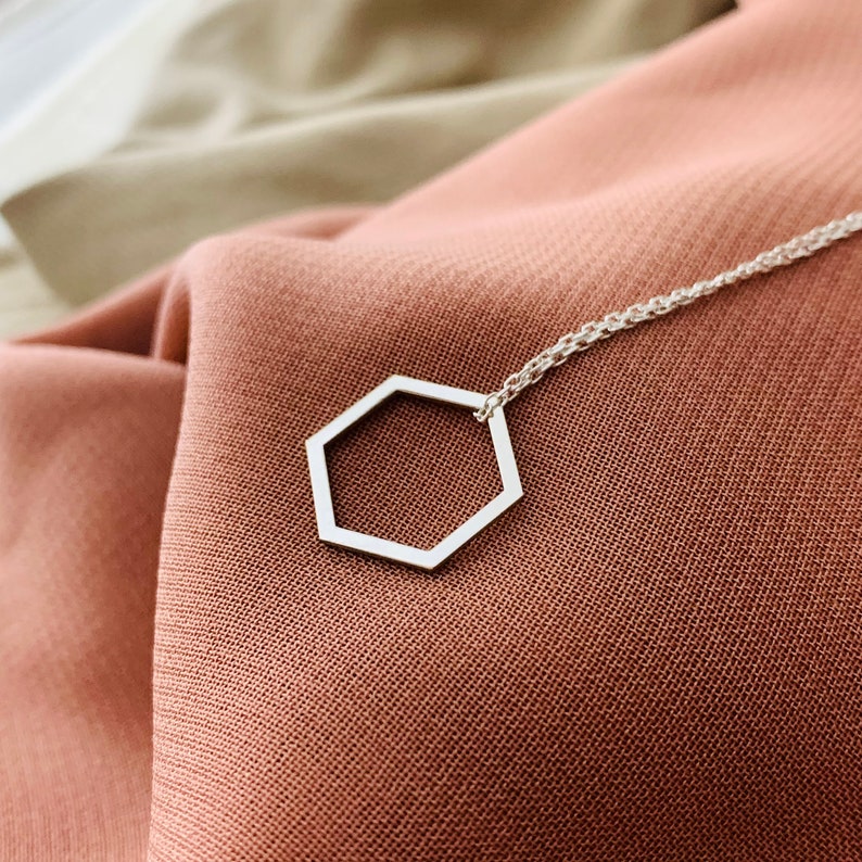 May include: A silver necklace with a hexagon pendant on a brown fabric background.