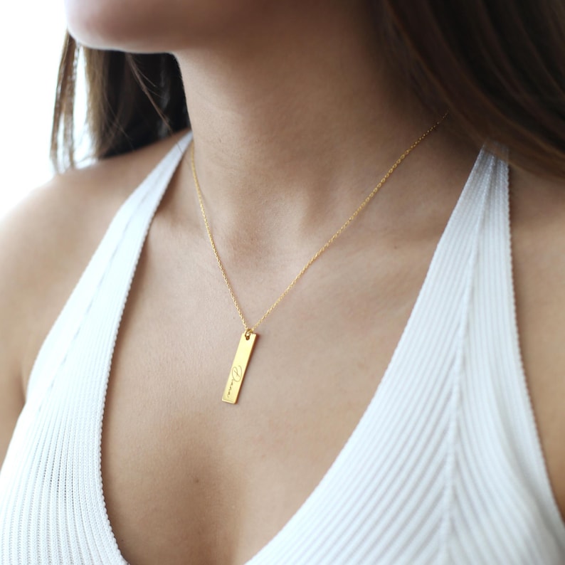 Puede incluir: Un collar de cadena de oro con un colgante rectangular delgado grabado con la palabra "Dreamer".