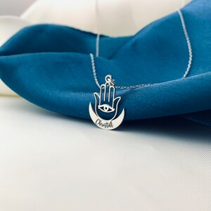 May include: Silver necklace with a hamsa hand charm and a crescent moon pendant. The pendant has an eye in the center and the name "Christal" engraved on the moon.