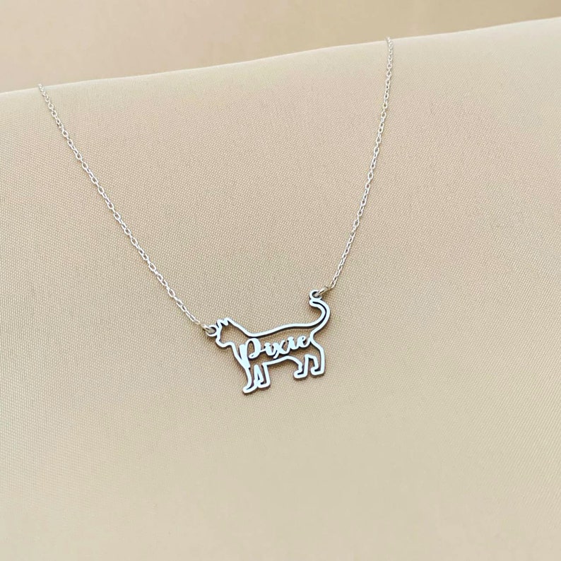 May include: Silver necklace featuring a cat silhouette pendant. The cat's name, "Pixie", is inscribed within the silhouette.