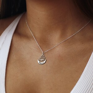 May include: A silver necklace with a hamsa hand charm with an eye in the palm and a crescent moon below. The name "Olivia" is engraved on the charm.