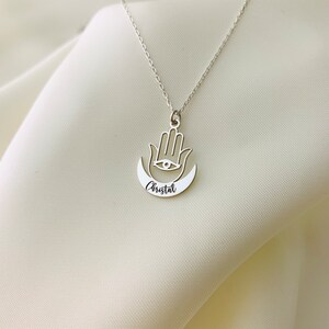 Pode incluir: Colar de prata com um pingente de m&atilde;o de hamsa e uma lua crescente com o nome "Cristal" gravado.