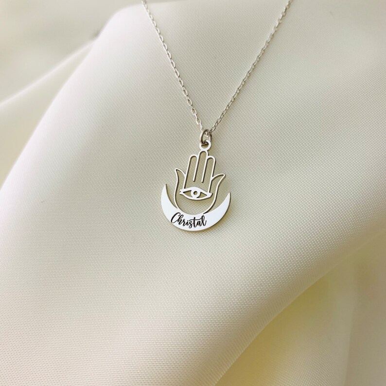 Pode incluir: Colar de prata com um pingente de m&atilde;o de hamsa e uma lua crescente com o nome "Cristal" gravado.