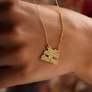May include: A gold-colored necklace with the phrase "fuck around & find out" in a stylized font. The pendant hangs from a delicate chain. The necklace is shown against a skin-toned background.