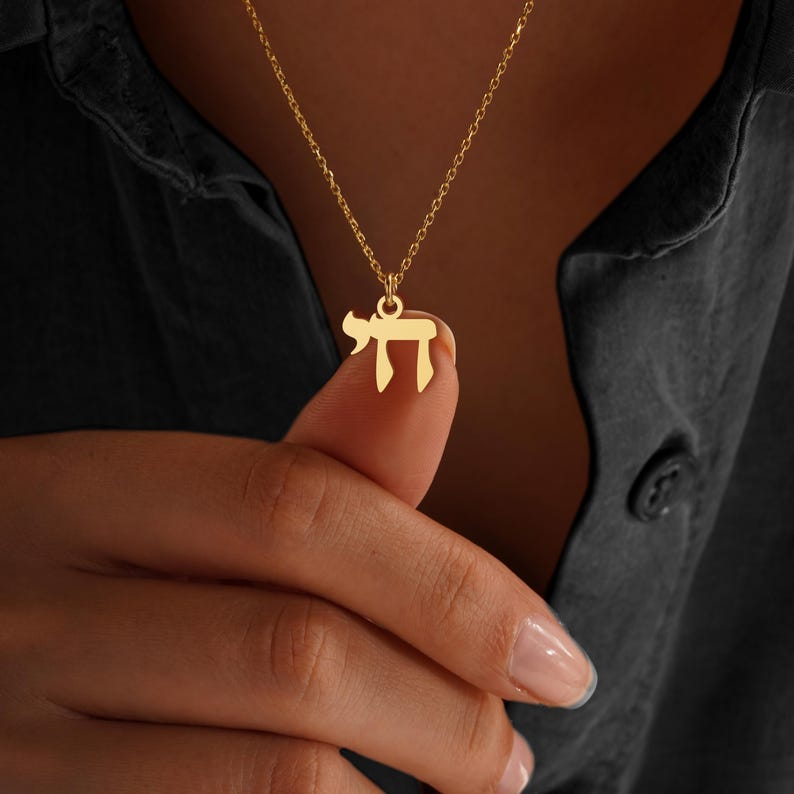 Peut inclure: Un collier dor&eacute; avec un pendentif en forme de la lettre h&eacute;bra&iuml;que 'Chai'. Le pendentif est suspendu &agrave; une cha&icirc;ne d&eacute;licate. Le collier est pr&eacute;sent&eacute; sur un fond sombre.