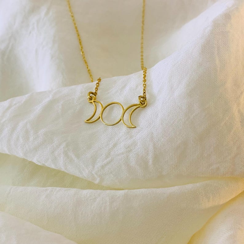 K&ouml;nnte beinhalten: Eine goldene Halskette mit einem dreifachen Mond-Anh&auml;nger. Der Anh&auml;nger ist ein schlichtes Design mit drei Mondsicheln.