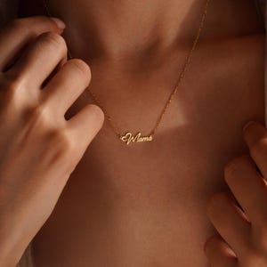 May include: A delicate gold necklace with the word "Mama" in cursive script. The necklace features a small heart detail and is worn around the neck. The chain is thin and the pendant is in a stylish font.