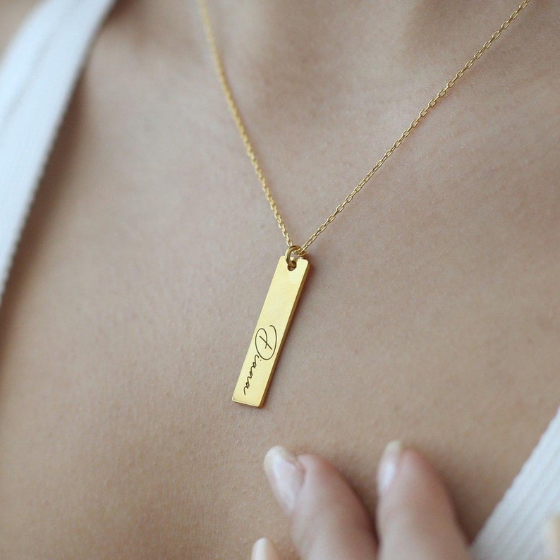 Puede incluir: Un collar de oro con un colgante de barra vertical grabado con el nombre "Diana" en escritura cursiva.