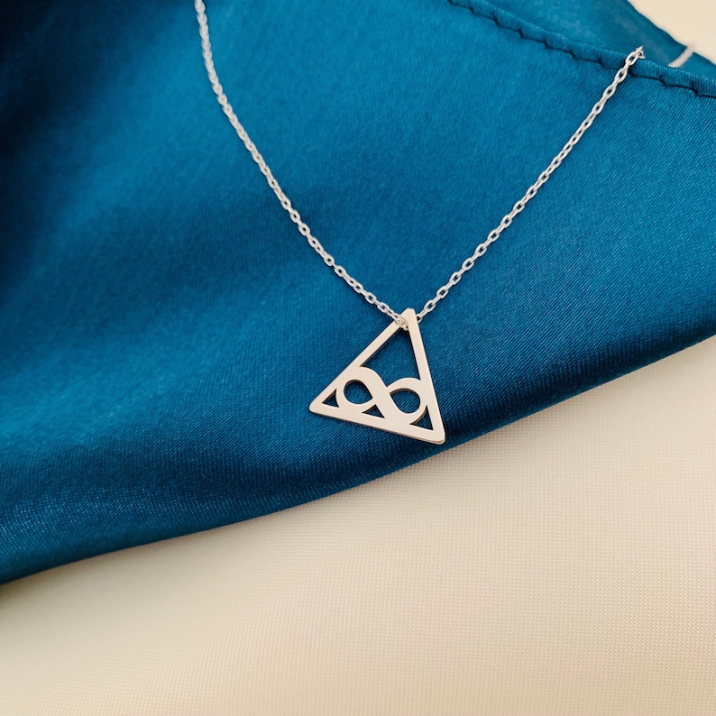 Pode incluir: Um colar de prata com um pingente triangular com um s&iacute;mbolo de infinito vazado. O pingente pende de uma corrente fina. O colar &eacute; exibido sobre um fundo de tecido azul-petr&oacute;leo.