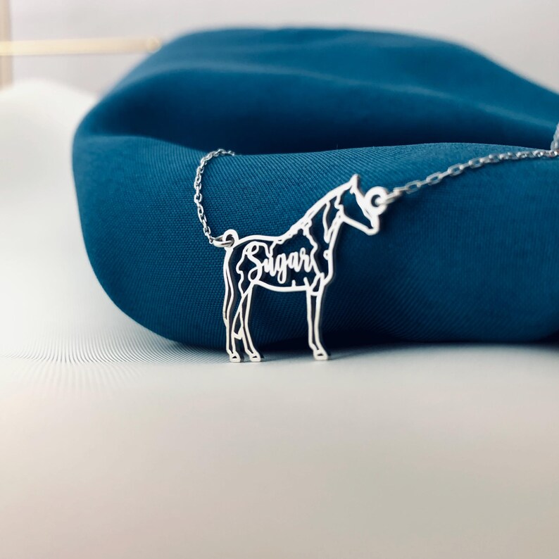 May include: A silver horse pendant necklace with the name "Sugar" inscribed on the horse's body. The necklace is displayed on a dark blue fabric background.