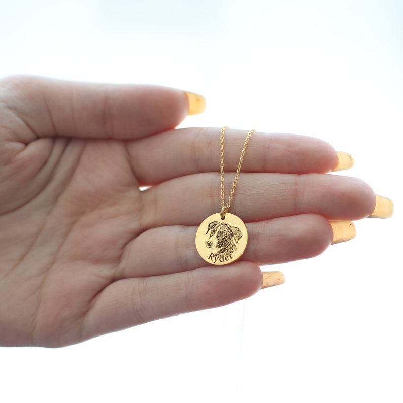 May include: A gold pendant necklace with a detailed engraved image of a dog's face. The round pendant has the dog's name, "Ryder," engraved below the image. The necklace chain is fine and gold-coloured.