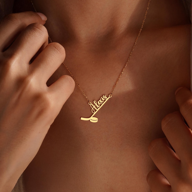 May include: A gold-colored necklace with the name "Alexis" in cursive script, accompanied by a hockey stick and puck design. The necklace is worn around the neck, with the chain visible. The pendant is a delicate piece of jewelry.