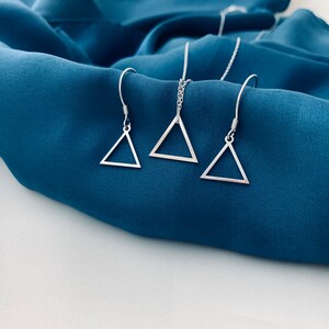 May include: A silver triangle necklace and matching earrings on a blue fabric background.