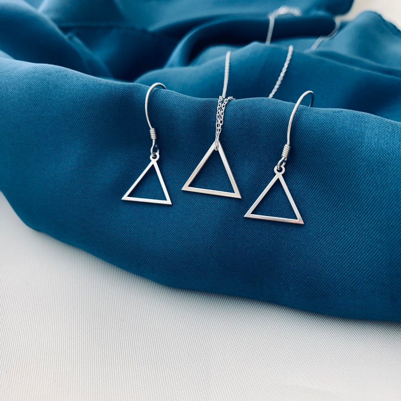 May include: A silver triangle necklace and matching earrings on a blue fabric background.