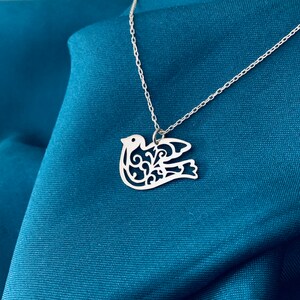 K&ouml;nnte beinhalten: Eine silberne Halskette mit einem filigranen, ausgeschnittenen Design einer Taube. Die Taube ist ein Symbol f&uuml;r Frieden und Hoffnung.