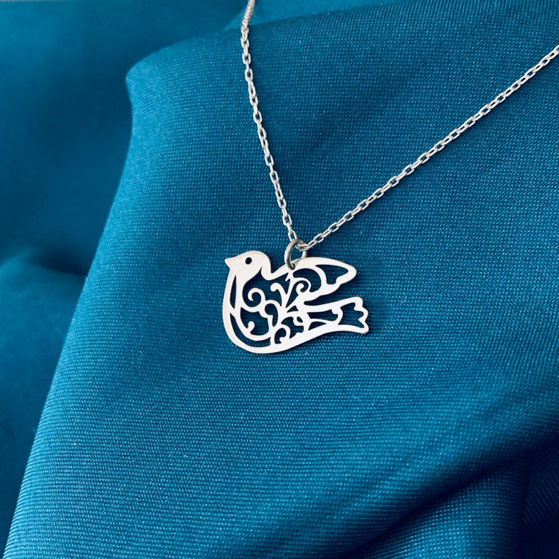 K&ouml;nnte beinhalten: Eine silberne Halskette mit einem filigranen, ausgeschnittenen Design einer Taube. Die Taube ist ein Symbol f&uuml;r Frieden und Hoffnung.