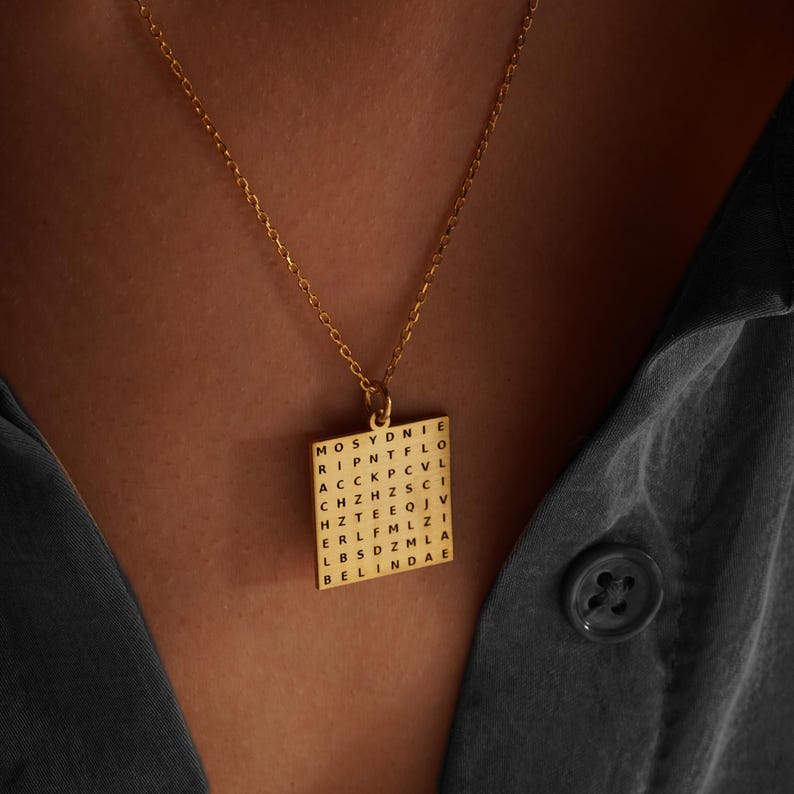 May include: A gold-colored pendant necklace with a square pendant featuring a word search design. The pendant is suspended from a delicate chain. The necklace is worn against a dark gray shirt.