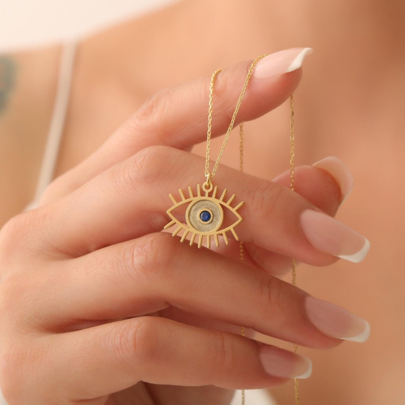 May include: Gold necklace featuring a blue evil eye pendant. The pendant is a detailed outline of an eye with a blue gemstone at its centre.