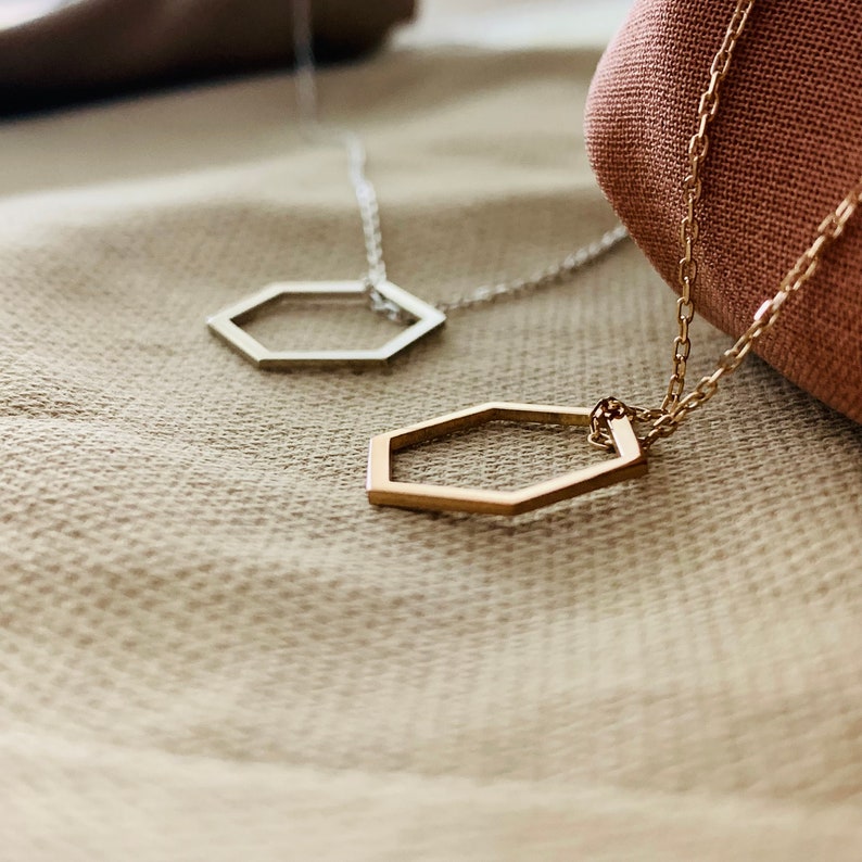 May include: Two gold necklaces with hexagon pendant charms. The necklaces are on a beige fabric background.