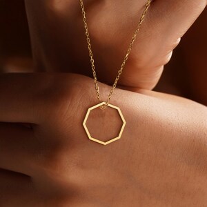Peut inclure: Un collier dor&eacute; avec un pendentif g&eacute;om&eacute;trique &agrave; huit c&ocirc;t&eacute;s. Le pendentif est suspendu &agrave; une fine cha&icirc;ne. L'image montre le collier sur un fond chaud et neutre.