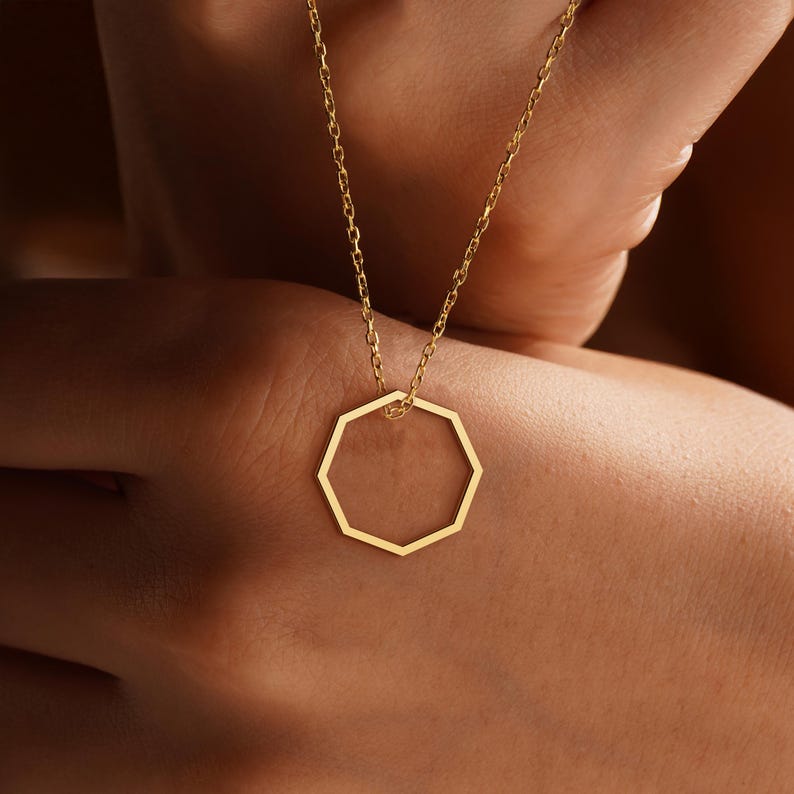 Peut inclure: Un collier dor&eacute; avec un pendentif g&eacute;om&eacute;trique &agrave; huit c&ocirc;t&eacute;s. Le pendentif est suspendu &agrave; une fine cha&icirc;ne. L'image montre le collier sur un fond chaud et neutre.