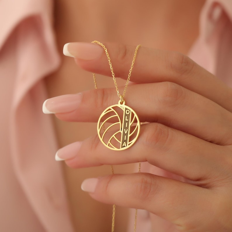 May include: A gold-coloured necklace with a volleyball pendant. The pendant is circular, displaying a volleyball design and the name "OLIVIA" vertically. The necklace is held by a person.