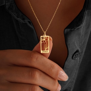 May include: A gold necklace with a rectangular pendant. The pendant has a floral design and the name "Jessica" engraved at the bottom. The necklace is shown against a dark background.