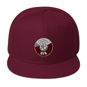 May include: A maroon flat-brimmed baseball cap with a circular embroidered patch. The patch features Arabic script, a soccer ball, and the letters "QFA" in white. The cap is a solid maroon color.