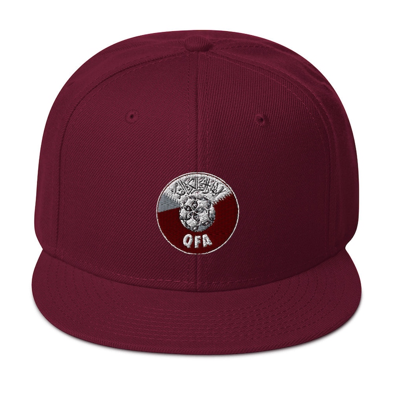 May include: A maroon flat-brimmed baseball cap with a circular embroidered patch. The patch features Arabic script, a soccer ball, and the letters "QFA" in white. The cap is a solid maroon color.