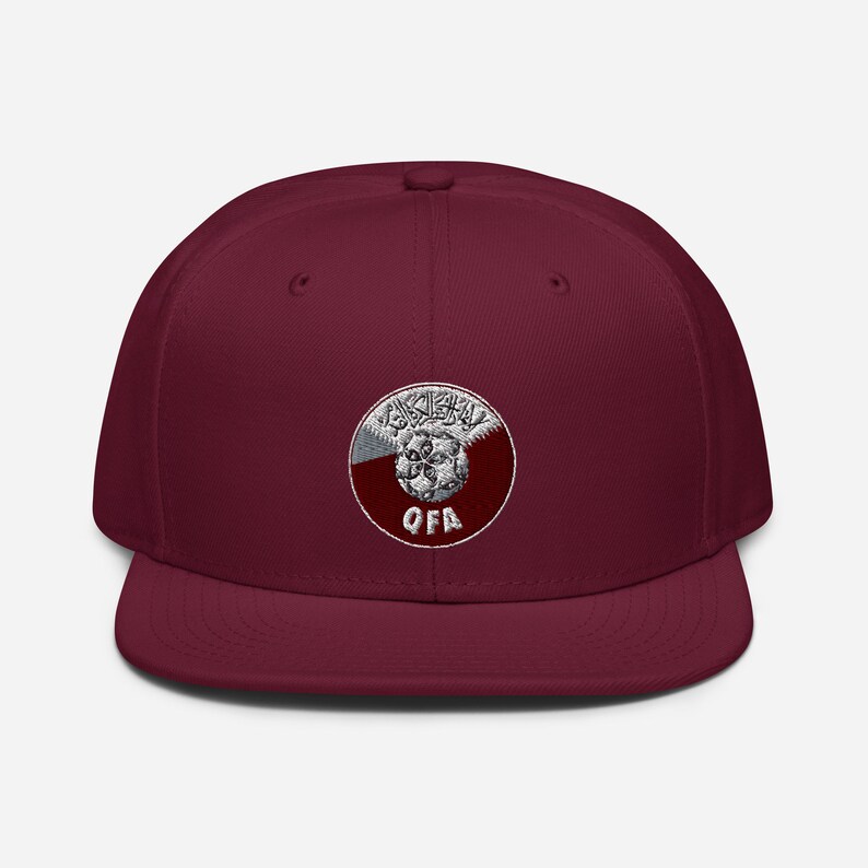 May include: A maroon flat-brim baseball cap featuring a circular emblem with Arabic script and the letters "QFA" in white. The emblem is set against a maroon and white background. The cap is a solid maroon color.