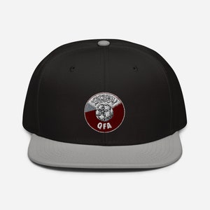 May include: A black and gray baseball cap with a flat brim. The cap features a circular emblem with Arabic script and the letters "QFA" in white. The emblem is set against a maroon and white background.