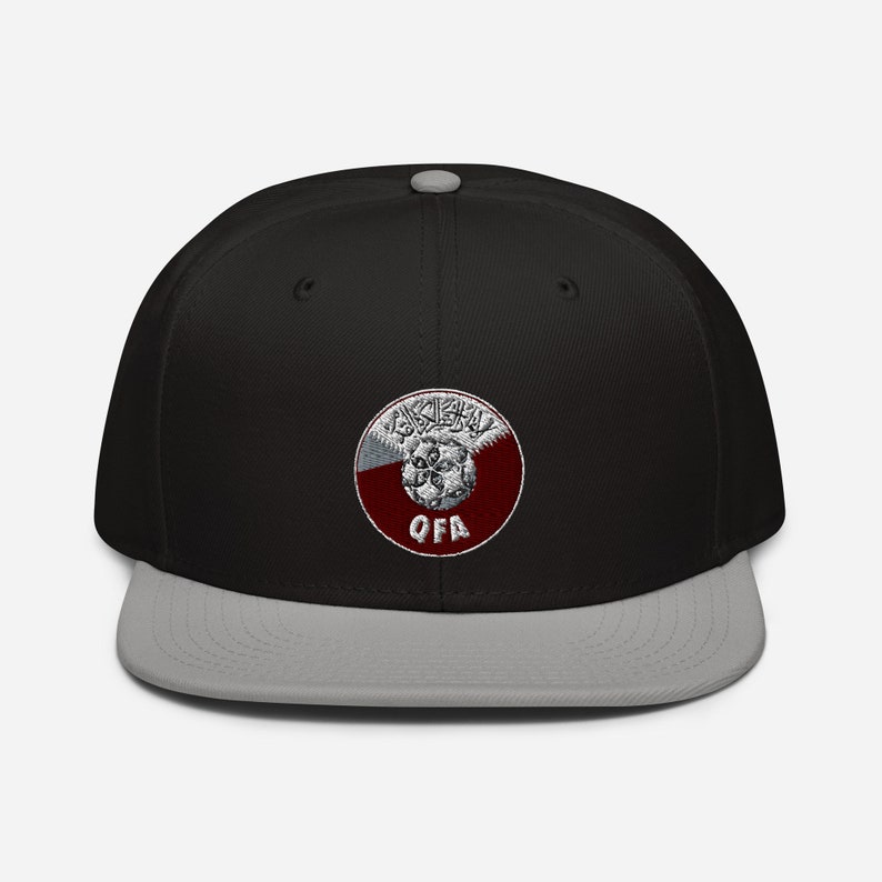 May include: A black and gray baseball cap with a flat brim. The cap features a circular emblem with Arabic script and the letters "QFA" in white. The emblem is set against a maroon and white background.