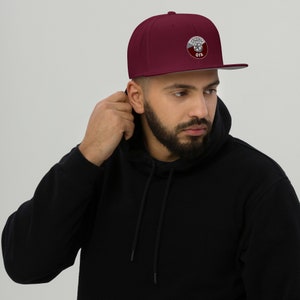 May include: A maroon flat-brimmed baseball cap with a white and maroon emblem. The emblem features a circular design with text. The cap has a structured fit and a gray brim. The person in the image is wearing a black hoodie.