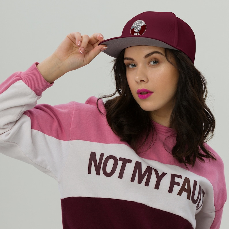 May include: A maroon flat-brimmed cap with a gray brim and a circular emblem. The emblem features a design with the letters "OFA". The cap is worn with a pink, white, and maroon color-block sweatshirt with the text "NOT MY FAULT".