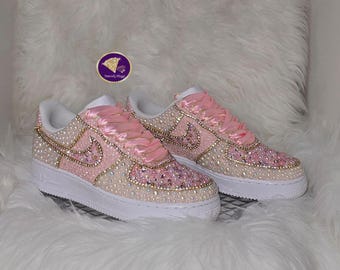 May include: Pink and cream-coloured trainers with rhinestone and gold accents. The shoes feature pink satin ribbon laces and a white sole. The Nike swoosh is outlined in gold. The trainers are embellished with rhinestones.