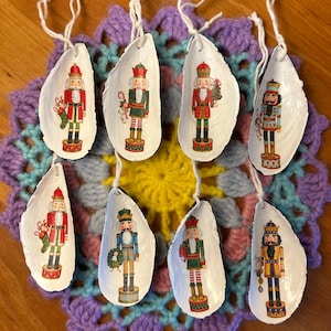 Op de afbeelding: Acht witte schelpenornamenten, elk met een kleurrijk notenkrakerontwerp. De ornamenten zijn opgehangen met wit touw en rusten op een veelkleurig gehaakt kleedje. De notenkrakers zijn afgebeeld in verschillende uniformen en poses.