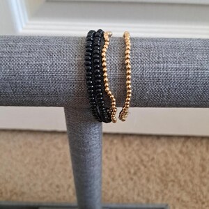 Pu&ograve; includere: Tre braccialetti di perline, uno nero e due dorati, esposti su un supporto in tessuto grigio.