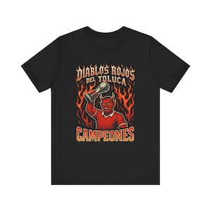 May include: Black t-shirt featuring a red devil holding a trophy, surrounded by flames. The text "DIABLOS ROJOS DEL TOLUCA" is arched above the devil, with "CAMPEONES" below. The design is a vintage-style illustration.